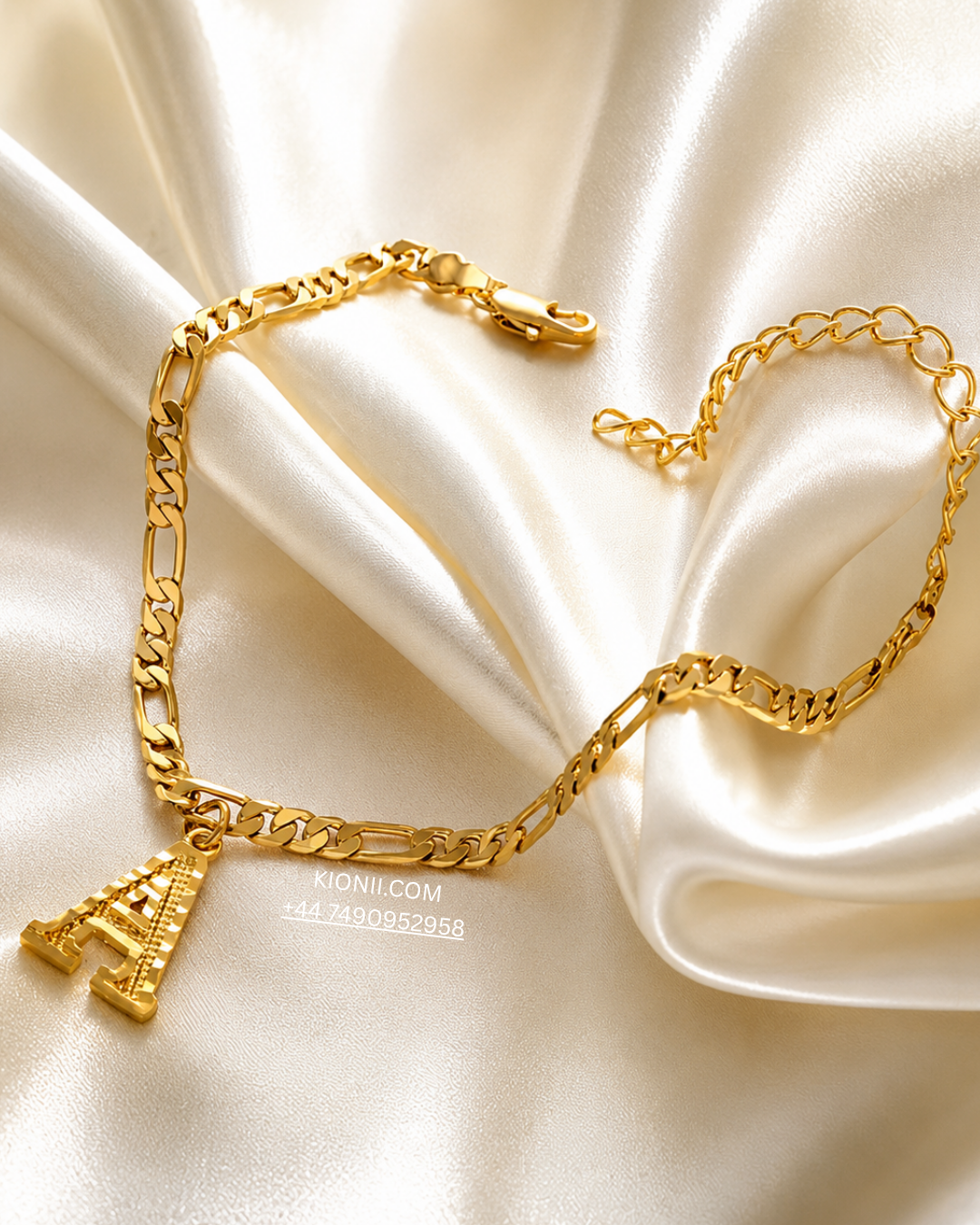 A gold-toned chain necklace with a small Eiffel Tower pendant, arranged on draped cream satin fabric. A small watermark reading MONIL.
