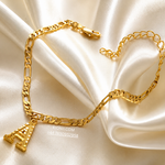 A gold-toned chain necklace with a small Eiffel Tower pendant, arranged on draped cream satin fabric. A small watermark reading MONIL.